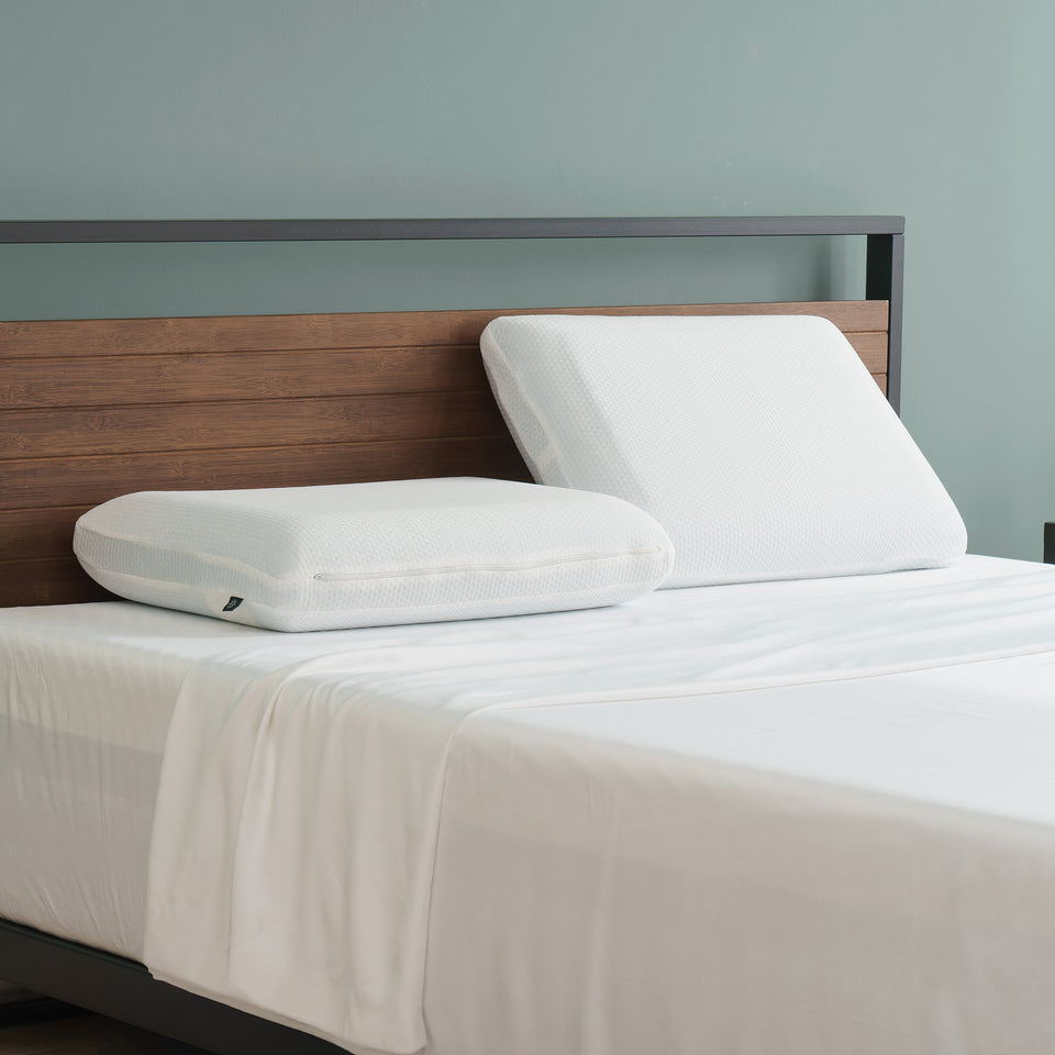 Two white memory foam pillows with breathable covers on neatly made bed, one laying flat and the other propped upright against wood headboard in a minimalist bedroom.