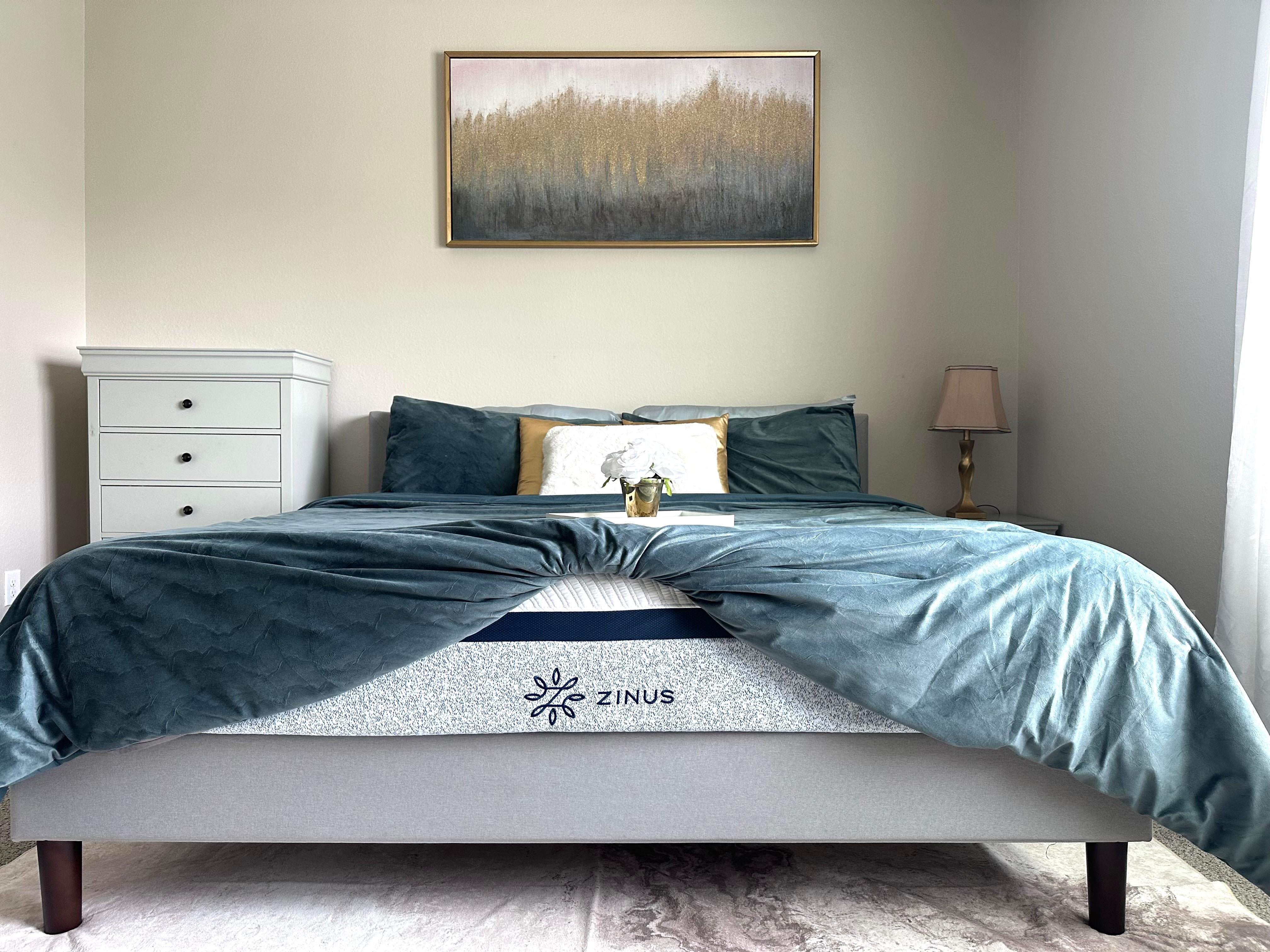 Zinus mattress with blue patterned side panel displayed on upholstered bed with teal bedding and accent pillows, styled in a bright bedroom with gold accents and a framed wall art.