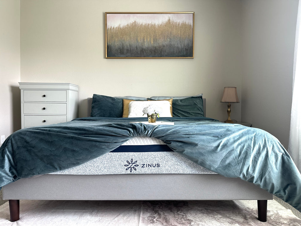 Zinus mattress with blue patterned side panel displayed on upholstered bed with teal bedding and accent pillows, styled in a bright bedroom with gold accents and a framed wall art.