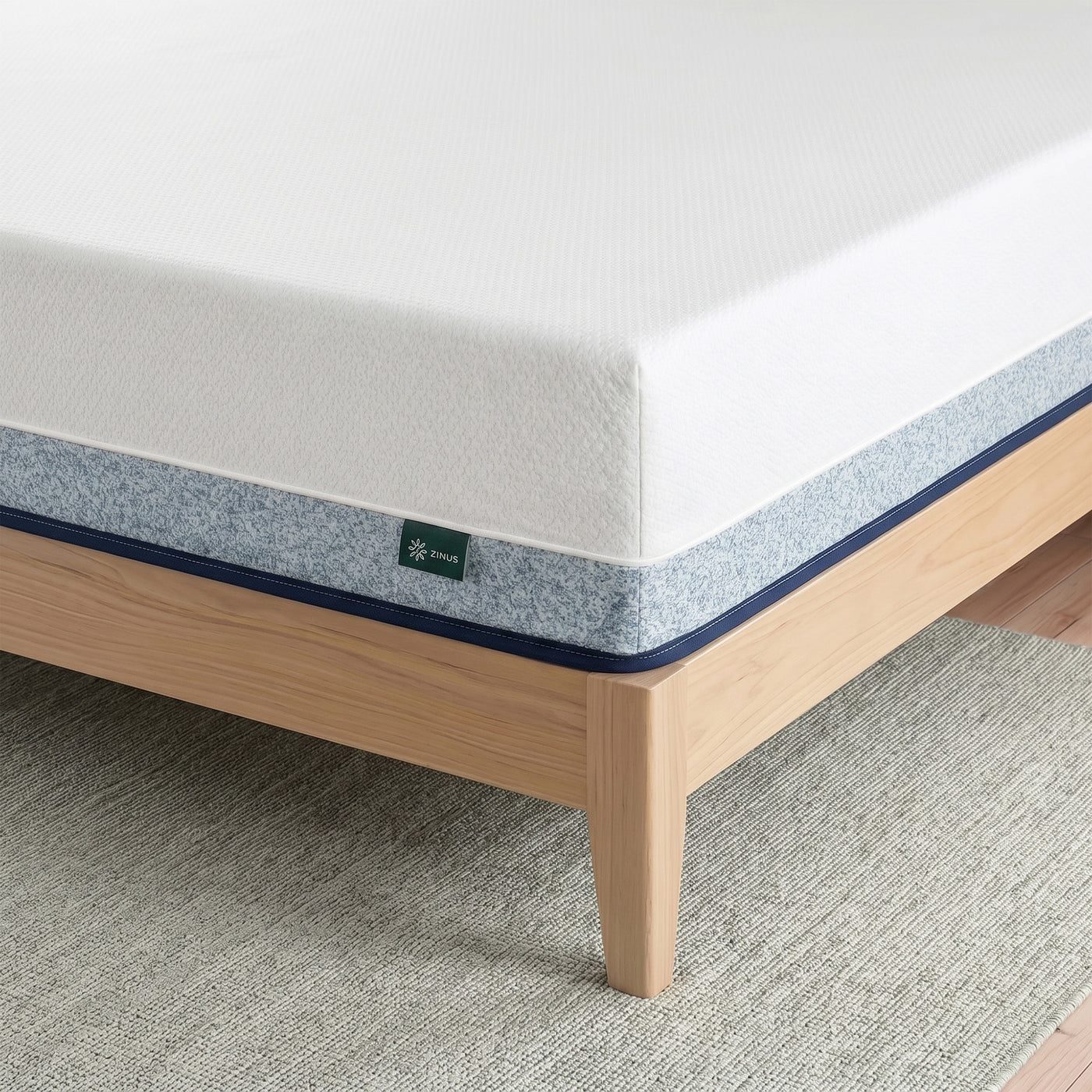 Zinus Original Green Tea Hybrid Mattress, Queen, close-up corner detail showing white knit top cover, gray textured side panel with navy piping, and Zinus logo tag, resting on light wood platform bed frame.